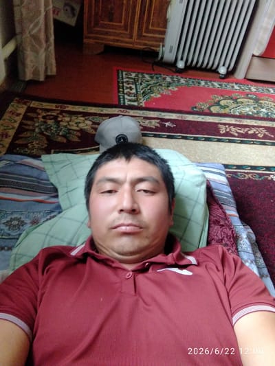 The image is a selfie taken from a close-up perspective, showing a man lying down. He is wearing a maroon polo shirt with white trim on the collar and sleeves. His head is resting on a light green pillow, and behind him, there appears to be a patterned blanket or bedding. In the background, several elements of an indoor setting are visible: a rug with intricate patterns, a wooden cabinet, and a white electric heater. The lighting suggests an indoor environment, potentially during the daytime. A timestamp in the lower right corner indicates "2026/6/22 12:00". The scene appears to be within a residential setting in Gul'cha, Kyrgyzstan.