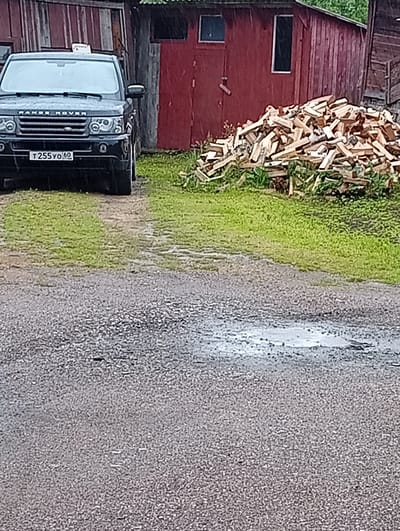 A black Range Rover Sport is parked on a gravel surface in front of a red wooden building with a pile of firewood to its right. The vehicle has a license plate that reads "T255YO60". The ground is wet, indicating it may be raining, and there is a small puddle in the foreground. The scene appears to be in a rural or semi-rural area, possibly in Ostrov-Zalit, Russia.