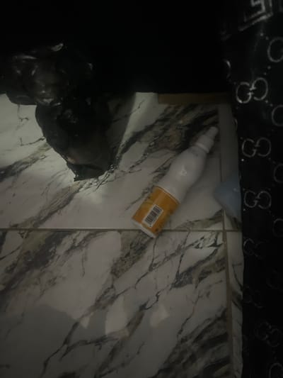 The image shows a white bottle with an orange label lying on a marbled floor. The bottle is cylindrical with a slightly curved body and a cap at the top. A barcode is visible on the orange label. To the left of the bottle, there is a dark, indistinct object, possibly a bag. Above the bottle and object, a dark fabric with a repeating pattern of interlocking "G" shapes is visible, suggesting it might be a garment or upholstery. The scene appears to be indoors, possibly a domestic setting, with the marbled floor providing a visual texture. The lighting is dim, casting shadows that obscure some details. There is no indication of time of day or weather. No people are visible in the frame. The text "1184001011" is visible on the barcode.