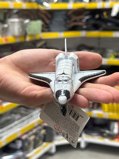 A person's hand holds a white toy space shuttle. The space shuttle is detailed, with black accents on the wings and a cockpit window. A white tag is attached to the shuttle. The background is blurred and shows shelves with products in a retail environment, possibly a supermarket or toy store, in the city of Ivanivka, Ukraine.