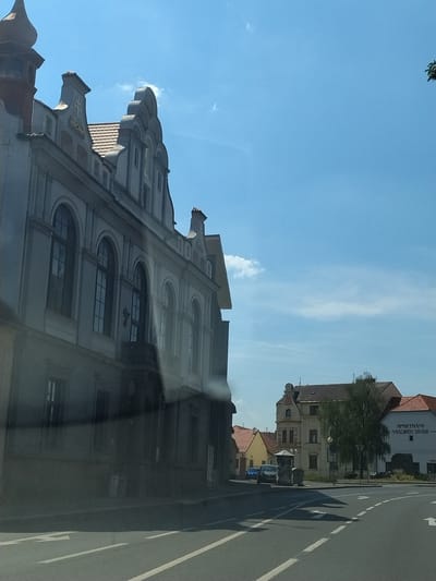 The image shows a street scene in Horažďovice, Czechia. On the left is a large, multi-story building with a grey facade and arched windows. The building has ornate architectural details on its roofline. To the right of this building, across the street, are several other buildings. One is a yellowish building with a red tile roof. Next to it is a taller, cream-colored building with a more modern appearance, featuring a sign that reads "APARTMÁNY NÁKOLÍ DVŮR". A large tree stands in front of this building. The street in the foreground is paved and has white lane markings. There are several cars visible parked or driving on the street. The sky is blue with a few scattered clouds. The image appears to be taken from a moving vehicle.