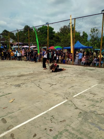 An outdoor event is depicted on a weathered concrete court with faded white lines and cracks, located in Tamansari, Indonesia. Under an overcast sky, two central figures are engaged in an activity. One individual stands upright, wearing dark trousers and a white shirt with a dark outer garment, holding a long, dark, whip-like object. Another individual is prostrate on the concrete, face down with arms extended, wearing dark clothing and a prominent headpiece adorned with long, dark, fibrous extensions. A crowd of approximately 50 to 70 spectators, including adults and children, lines the perimeter behind a black mesh net supported by poles. Some spectators are seated, others stand, with several gathered under temporary shelters and colorful umbrellas (blue, red, green). Two tall, narrow flags, one green and one gold, are attached to the net poles. Lush green trees and foliage form the background behind the spectator area and temporary structures. A single light-colored sandal rests on the concrete court in the left foreground.