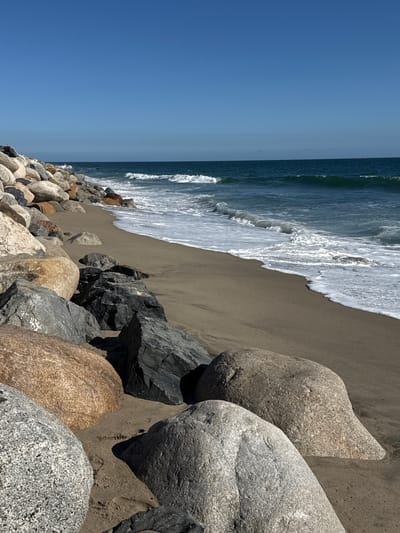 This is a photograph of a rocky beach in Malibu, California, under a clear blue sky. The foreground is dominated by large, weathered boulders of various sizes and colors, ranging from light grey to tan and dark grey. The boulders are partially submerged in dark sand. A gentle wave is washing ashore, creating a foamy white line along the edge of the water. The ocean is a deep blue-green, with several small waves visible in the distance. The horizon is sharply defined by the meeting of the sea and sky. No people or animals are present in the image. The scene is tranquil and natural.