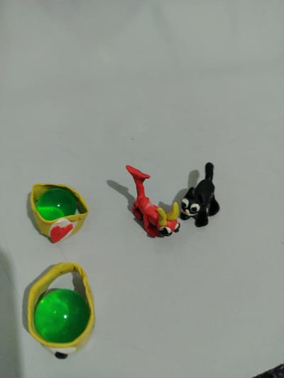 The image depicts two small, handmade clay figures on a light gray, textured surface. On the left are two yellow, cup-like objects, each containing a translucent green sphere. The upper cup has a red heart shape affixed to its side. In the center and to the right, a red, dragon-like creature with yellow horns is positioned facing a small black cat with wide white eyes. The cat is looking towards the dragon. The scene is simple, with no discernible background details other than the plain surface. The lighting suggests an indoor setting, possibly with overhead light, casting soft shadows from the figures onto the surface. There is no visible text. The overall impression is of a playful, handcrafted display. The mention of "Şamaldı-Say, Kyrgyzstan" provides a geographical context for the image, suggesting this might be a scene from that region, though no specific location cues are visually present in the image itself.