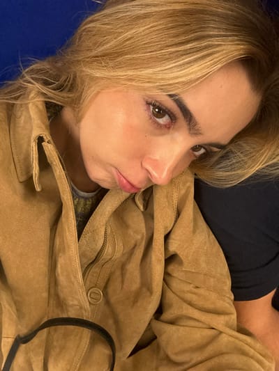 A female individual with blonde hair and fair skin is depicted in a close-up, head-and-shoulders portrait. Her head is tilted to the left, resting against an unseen dark blue or black surface, with her gaze directed forward towards the viewer. She has brown eyes, defined eyebrows, and appears to be wearing mascara and a light lip gloss on her slightly pouting lips. She is dressed in a tan or light brown suede-like jacket, featuring a collar, a visible zipper running down the front, and a circular snap button closure on the lower front left. A segment of a patterned garment, possibly a green, yellow, and grey sweater, is visible beneath the open jacket collar. The upper left background reveals a solid blue surface. A black strap is visible in the lower left foreground. The specific location within the city of Best, Netherlands, or any other location, cannot be determined from the visual information provided in the image.