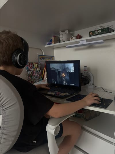 A young individual, appearing male, with light brown hair, is seated in a grey and white ergonomic chair at a white desk in an indoor setting located in Krasnaya Zarya, Russia. The individual wears black over-ear headphones with a white lightning bolt logo and a black t-shirt with grey side panels, along with black shorts. They are actively playing a first-person shooter video game on a black laptop, which displays an urban combat scene with explosions, smoke, and an aiming reticle. The individual's left hand rests on the laptop keyboard, while the right hand operates a black mouse on a black mousepad featuring a "SONNEN" logo and gold-toned patterns. The laptop keyboard is decorated with various stickers, including several depicting anime-style eyes.
Above the desk, a white floating shelf holds a white ceramic piggy bank, a small red container, a white decorative object, and a green box labeled "MEMO 50." A white LED strip light is mounted beneath this shelf. To the left of the laptop are stacked books and a board covered with colorful alphabet stickers. To the right, a white adjustable device, resembling a small microscope or desk lamp, is visible, along with associated black wiring. Below the desk, a camouflage-patterned cylindrical object is partially seen between the chair armrest and white desk drawers.