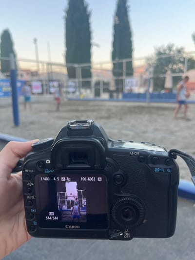 A person's hand holds a black Canon DSLR camera, focused on a beach volleyball court. The camera's LCD screen displays a photo of three people on the court, two in the foreground and one further back. The foreground figures are wearing white t-shirts and blue shorts, with one reaching up to hit a volleyball. The background is slightly blurred, showing more of the court, nets, trees, and a clear sky. The environment appears to be outdoors, likely a beach or a sandy court in a location such as Gošići, Montenegro, given the presence of tall, slender trees and the architectural style of the distant structures. The overall lighting suggests it is late afternoon or early evening.