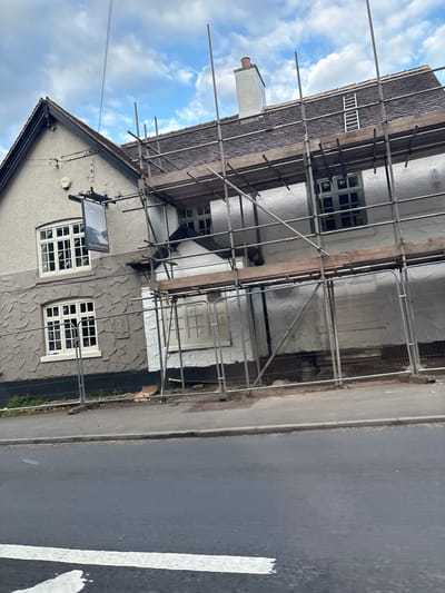 The image shows a building under renovation in Wistaston, United Kingdom. The building, likely a public house as indicated by a sign reading "The Rising Sun", is covered in scaffolding and protective sheeting. The facade is a mixture of textured gray render and white painted sections. There are several windows visible, some with multiple panes and green frames. A chimney stack is visible on the roof, which is tiled. The scene is situated alongside a road, with a pavement and a safety fence in front of the building. The sky is partly cloudy.