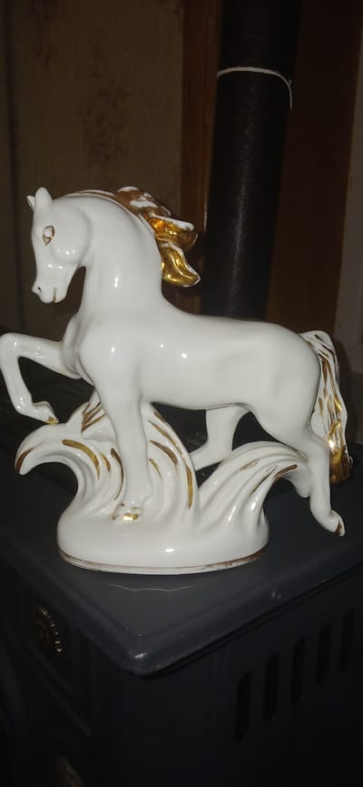 A white porcelain horse figurine with gold accents is displayed on a dark gray, ribbed surface. The figurine depicts a horse in motion, with its front legs raised and its mane and tail adorned with gold. The horse stands on a base that resembles waves, also decorated with gold trim. The background is blurred, showing a textured wall in a light brown hue and a dark cylindrical object. The figurine is illuminated by soft light, casting subtle shadows. The surface it rests upon appears to be the top of a stove or a similar appliance, with vents visible on its side. The scene suggests an indoor setting, possibly a home environment. There is no indication of Ganja City, Azerbaijan.