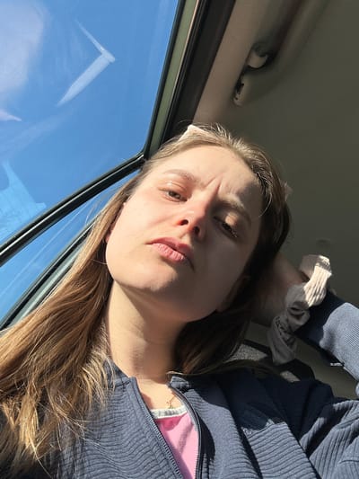 A young woman is shown in a close-up, angled selfie taken inside a vehicle. The visible background through the car window is a clear blue sky. She is wearing a dark blue, possibly corduroy, zip-up jacket over a pink top. A light-colored scrunchie appears near the top of the frame, seemingly holding up her hair. The location is unknown, but possibly Denmark, based on the prompt.