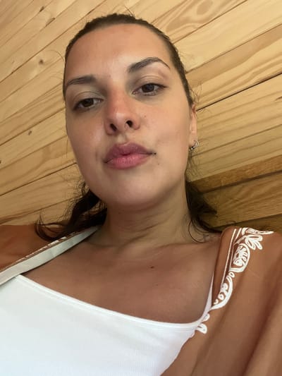 This is a close-up, sideways portrait of a young woman. She has dark hair and is wearing a white tank top. The background is made of light-colored wooden planks. A brown fabric with white intricate patterns is visible in the upper right corner, draped over her shoulder. The woman's face is the primary focus, with her eyes looking towards the camera and her lips slightly parted. Her skin appears natural and dewy. A small silver hoop earring is visible in her left ear. The lighting suggests it could be daytime. The overall impression is casual and intimate.