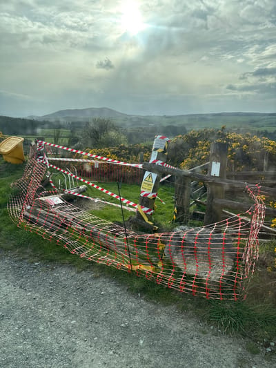 The image shows a rural scene in Shrewsbury, United Kingdom. A damaged wooden gate is heavily barricaded with red and white striped hazard tape and orange plastic netting. Several warning signs are visible, including one with a lightning bolt symbol indicating electrical danger. The gate appears to lead to a grassy field or hillside with yellow flowering bushes. The background features rolling hills and a cloudy sky. The foreground shows a portion of a gravel or dirt road. A yellow bin is partially visible on the lower left. The scene suggests a potential hazard or restricted access area.