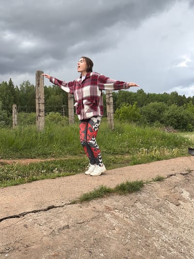 A young person stands with arms outstretched on a concrete edge in Nizhnii Vishur, Russia. The person wears a plaid shirt, patterned pants, and white sneakers. The background features green grass, trees, a cloudy sky, and a concrete fence.
