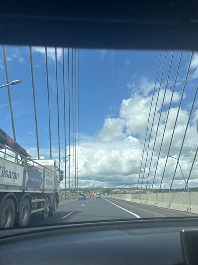 This image, taken from inside a vehicle, shows a view of a bridge and the sky. The perspective is angled, suggesting the photo was taken from a side window. The foreground is dominated by dark, blurred elements, possibly the interior of the vehicle. Stretching across the frame are numerous parallel suspension cables, a defining feature of the bridge's structure.
Beyond the cables, a bright blue sky with fluffy white clouds is visible. The sky takes up a significant portion of the image, creating a sense of openness. A highway runs horizontally across the middle ground, with a white dividing line visible. Several vehicles, including a truck and a car, are driving on the highway. The truck has a white trailer with blue and white text and a logo. The text "Kilsaran" is clearly visible on the side of the trailer, along with "www.kilsaran.ie".
Further in the distance, a treeline and buildings can be seen along the horizon, suggesting a populated area beyond the bridge. Lampposts line the side of the highway. The lighting indicates it is daytime, likely with good weather due to the clear sky and bright clouds. The overall impression is a snapshot of travel on a bridge on a clear day.