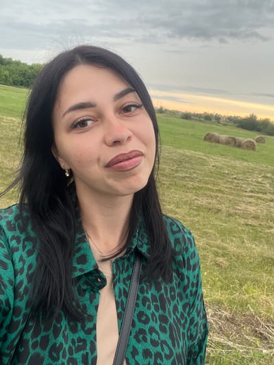 A medium shot captures a woman with long, dark hair and medium skin tone from the chest up, facing the camera directly. She is outdoors in a grassy field with scattered hay bales in the background, under a cloudy sky with a hint of sunset colors on the horizon. The woman wears a green and black leopard-print button-down shirt over a beige tank top. She has on light-colored earrings and a delicate necklace. Her makeup includes brown lipstick and mascara. To her right, a dark strap, possibly from a bag, is visible across her chest. The setting appears to be a rural area, potentially on the outskirts of Istobnoye, Russia, given the natural landscape.