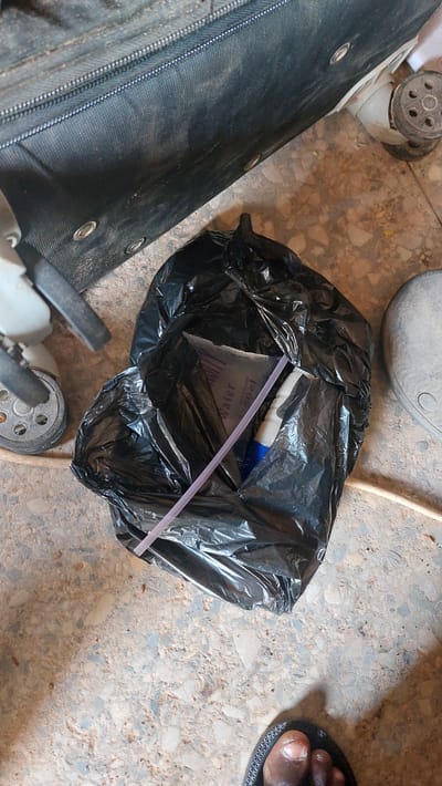 A black trash bag lies on a terrazzo floor in New Nyanya, Nigeria. Inside the bag, a purple straw is visible, along with a small, clear pouch containing liquid labeled "Water 50cl". A white bottle with a blue cap is also partially visible. To the left of the trash bag, a black suitcase is partially visible. To the right of the trash bag, a person's foot wearing a black sandal is in the lower right corner of the frame. The environment appears to be indoors, possibly a storage area or a hallway. The lighting is overhead, casting shadows on the floor.