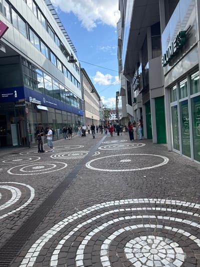 This image captures a wide-angle view of a bustling pedestrian street in the city center of Darmstadt, Germany, under a clear sky with scattered clouds.

On the left, a modern multi-story building with a facade primarily composed of large, reflective windows dominates the upper half of the frame. The ground floor of this building features a prominent blue band with white lettering that reads "Wissenschaftsstadt Darmstadt" and "Rathaus" (City Hall), identifying the location. Below this, partially obscured, are other informational signs. Near the building's entrance, a dark green cylindrical trash receptacle stands next to a pole displaying a red "Einfahrt freihalten" (Keep entrance clear) sign, a "Radfahrer absteigen" (Cyclists dismount) sign, and an arrow pointing towards "Centralstation."

Standing on the cobblestone pavement in front of the Rathaus are three individuals. A woman on the left wears a black top, patterned black tights, and platform shoes, holding a small black bag. She is engaged with a man next to her, who has dark hair and wears a dark t-shirt and dark trousers, looking at his phone. To his right, another man in a white t-shirt, light blue shorts, and white sneakers is also absorbed in his phone. Further down the street, multiple pedestrians are visible, some walking individually, others in small groups, including one person pushing a stroller.

The right side of the street features commercial buildings. A sign with prominent green lettering on a building facade reads "BLUME 2000," accompanied by a stylized floral logo. Below this, reflecting in the glass of an adjacent building, the word "LUISEN CENTER" is faintly discernible. The storefronts on the immediate right are characterized by tall, green-tinted glass panels, some showing reflections of faint QR codes and text like "7D AY." Three young women are gathered outside one of these storefronts: one in a white top and dark pants, another in a light brown top and dark pants, and a third in a light blue top and light jeans.

The street itself is paved with dark gray cobblestones arranged in a fan-like pattern. Integrated into this paving are distinctive white circular patterns, appearing as concentric rings with a central white rosette. A long, dark metal drainage grate runs longitudinally down the center of the cobblestone street. The bright blue sky with fluffy white clouds suggests fair weather conditions.