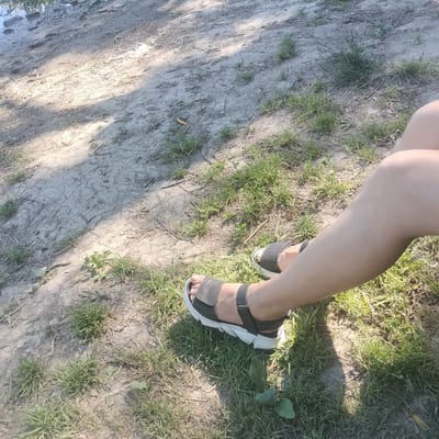 The image displays a person's legs and feet, clad in black sandals with white soles, resting on a patch of dry, sandy ground interspersed with patches of green grass. The ground shows signs of drought, with visible cracks. Sunlight casts dappled shadows across the terrain, suggesting an outdoor, possibly park-like setting. The background is a mix of sandy soil and vegetation. There is no indication of the city of Sheptytskyi, Ukraine, in the image.
