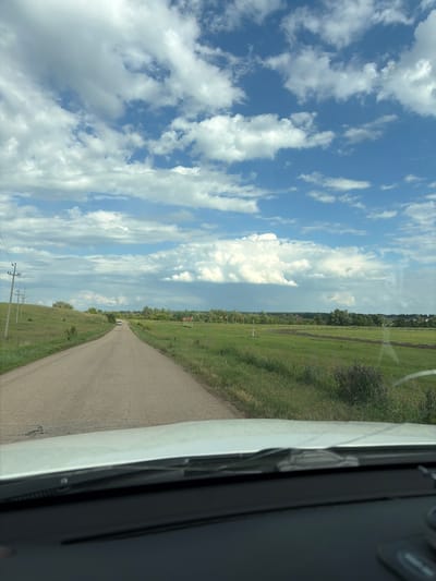 A white car is driving on a dirt road through a rural landscape. The road is lined with green grass and fields on either side. In the distance, there are trees and a cloudy sky. There are power poles along the left side of the road. The image appears to be taken from inside the car, looking forward.
