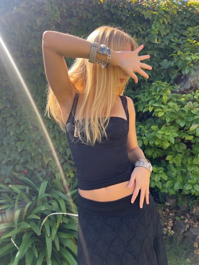 A lone individual, appearing female, is depicted standing outdoors against a backdrop of dense green foliage in Quilmes Oeste, Argentina. The person has long, straight, light-blonde hair. Their face is partially obscured by their right hand, which is raised and splayed, covering the eyes, consistent with shielding from bright sunlight. A strong lens flare is visible in the upper-left quadrant of the image.
The individual wears a form-fitting black sleeveless top featuring a lace-trimmed bustline, which exposes a section of the midriff. Below this, they wear a long, black skirt with a distinct textured or lace pattern. Both wrists are accessorized with multiple silver-toned bracelets. The right wrist displays at least two distinct cuffs, one with a textured surface and another with multiple parallel ridges. The left wrist features a single wide, intricately patterned silver-toned cuff.
The background consists of a layered natural environment: a dark green, dense wall of climbing ivy or similar foliage occupies the upper left, while brighter green shrubbery or bushes are visible across the middle and right-hand side. The ground is partially visible in the lower right, appearing as dark soil or organic matter. The scene is brightly lit by natural sunlight, originating from the upper-left, indicated by the prominent flare. No other individuals or objects are observed in the frame.