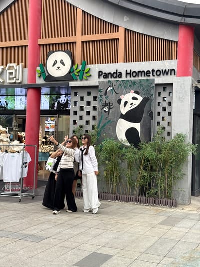 The scene is a commercial storefront identified as “Panda Hometown,” possibly a souvenir or theme shop, located in what appears to be Chenghua District, China, given the signage featuring pandas and Chinese characters. Three women are posing in front of the store, possibly taking photos. One woman is wearing a white shirt and white pants, while the other two wear striped shirts and black pants. A merchandise display with clothes, hats, and water bottles is situated in the foreground. The store’s facade includes panda-themed decorations, a painting of a reclining panda, and a green bamboo-like feature. A menu board or screen displaying panda images and food items is visible inside the store.