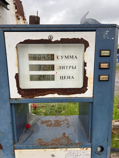 A close-up, eye-level, outdoor shot shows a weathered, blue, metal fuel dispenser, likely located in Kopeysk, Russia, given the Cyrillic text. The dispenser's front panel features a digital display showing three lines of information: "804.50" labeled "СУММА" (SUMMA - amount), "32.18" labeled "ЛИТРЫ" (LITRY - liters), and "25.00" labeled "ЦЕНА" (TSENA - price). The display is framed by a rusty, white border. To the right of the display are three rectangular buttons with Cyrillic labels: "ПУСК" (PUSK - start) in green, "СТОП" (STOP - stop) in red, and "СБРОС" (SBROS - reset) in grey. The dispenser is positioned in front of an overcast sky and a background of green foliage, suggesting an outdoor location. Below the digital display, there is a shelf holding a grey plastic funnel with a red cap and a wire handle. The overall condition of the dispenser indicates age and exposure to the elements, with visible rust and chipped paint.