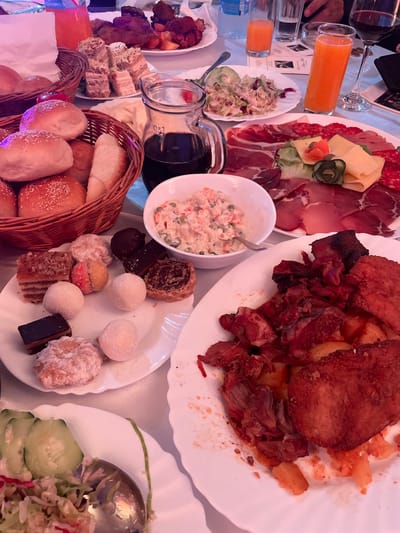 The image depicts a table laden with food, likely in the city of Беране, Montenegro. A variety of dishes are visible, including a plate with cured meats and cheese, a plate of fried meat and potatoes, a bowl of Russian salad, a pitcher of dark liquid (likely wine or juice), a basket of bread rolls, a plate of assorted pastries, and a salad with cucumbers and cabbage. Two glasses of orange juice are also visible. The scene appears to be a communal meal or a buffet setting. No people are fully visible, only hands and arms.