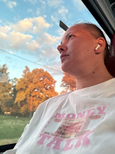 A close-up, rotated shot captures a young person, possibly female, with their hair pulled back, looking downwards or to the side with a neutral, perhaps pensive, expression. They are wearing a white t-shirt adorned with prominent pink text. Visible on the shirt are the words "MONEY TALK" in a stylized font, accompanied by smaller numbers like "5000," suggesting a design reminiscent of currency. A white earbud is clearly visible in their right ear.
The subject appears to be seated next to a window, likely in a moving vehicle, indicated by a dark interior element in the upper left and subtle reflections. The background outside the window reveals a light blue sky with scattered white and grey clouds. Below the sky, several deciduous trees display vibrant golden-orange foliage, suggesting either late autumn or the warm, soft light of late afternoon or early evening, possibly during sunset. A blurred patch of green, likely grass or ground cover, is visible beneath the trees. The overall lighting creates a serene atmosphere during what seems to be a journey through a rural or suburban landscape near Базарово, Russia.
