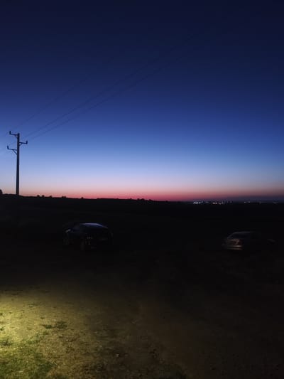 The image depicts a twilight scene in Ravadinovo, Bulgaria, with a clear, deep blue sky transitioning to a faint pink and orange hue at the horizon. A power line runs horizontally across the upper portion of the frame, supported by a utility pole on the left. The ground in the foreground is illuminated by a bright, artificial light source, casting a yellow glow on the rough terrain, which appears to be a dirt track with patches of dry grass. In the middle ground, silhouetted against the darkening landscape, are two cars. One is on the left, a darker vehicle, and another is on the right, a lighter-colored sedan. The background is a dark silhouette of rolling hills, with distant lights suggesting a town or village. The overall atmosphere is serene and quiet, characteristic of dusk.