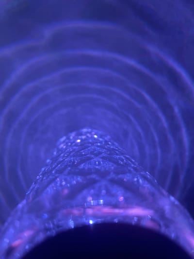 This abstract image, located in Dmitriyevskaya, Russia, displays a close-up view dominated by shades of purple and blue light. The central foreground features a prominent, elongated, translucent object, likely glass or crystal, extending towards the viewer. This object possesses a heavily textured or faceted surface, exhibiting numerous small, sparkling points of light, indicating refraction and reflection. The pattern on its surface appears intricate and repetitive. In the background, soft, concentric or arcing wave-like patterns of purple light are visible, out of sharp focus, suggesting further light refractions or reflections within the environment. The lower portion of the main object curves downwards, leading to an obscured dark area at the bottom of the frame. There are no discernible people, specific actions, or conventional environmental elements beyond the interplay of light and patterned surfaces.