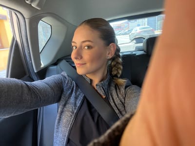The image depicts a female individual seated in the rear passenger compartment of a vehicle. She has light-colored eyes and her hair is styled in a single braid extending over her left shoulder. A small, round, silver-colored stud earring is visible in her left ear. She is wearing a dark grey long-sleeved athletic-style top with a zipper, over a black garment, and is secured by a black vehicle seatbelt diagonally across her chest. Her right arm is extended towards the camera, with a significant portion of her hand and fingers appearing as a large, out-of-focus orange-pink blur in the right foreground of the frame, indicating she is holding the camera for a selfie. She is looking directly at the camera with a slight smile.
The vehicle's interior features black seating and grey plastic trim around the windows. Through the open window on the left side of the vehicle, a portion of a light yellow-colored building facade is visible. Through the rear window, an urban or commercial street environment is discernible, including multiple parked or moving vehicles, prominently a white sedan in the mid-ground. In the background, a large commercial building with red and white signage, partially visible (possibly reading "MEGA" or similar), suggests a retail or industrial area. The scene is located in the city of Camargo, Spain.