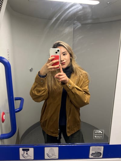 A female individual is depicted in a mirror selfie taken inside an enclosed space, likely a public restroom or small cubicle, characterized by light grey walls and a white ceiling. A horizontal fluorescent light fixture is visible at the top. On the left, a blue U-shaped grab bar is affixed to the wall, with a circular red push-button positioned beneath it. The bottom edge of the mirror features a blue frame displaying white pictograms for a soap dispenser, hand washing, and hand drying.
The subject is a woman with long, straight light-brown hair, wearing a dark-colored turtleneck top beneath an open, tan-colored suede or faux-suede jacket. She holds a smartphone, equipped with a clear case and a red "pop-it" style accessory, in her left hand to capture her reflection. Her right hand is raised, with the index finger extended and slightly bent, pointing towards her chin area. She is looking directly into the camera lens. The specific location within Mandello del Lario, Italy, cannot be determined from the visual evidence provided.