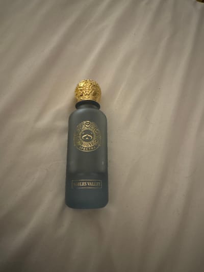 A dark matte perfume bottle with a gold ornate cap stands upright on a textured, light-colored surface, likely fabric. The bottle has a circular emblem in gold with the word "SPECTRA" in a smaller font below it. Near the bottom of the bottle, the words "NOBLES VALLEY" are inscribed in a gold rectangular box. The background is a soft, wrinkled fabric. The image appears to be a product photograph, likely taken indoors. There is no indication of the city of Sharjah, United Arab Emirates in the image itself or its context.