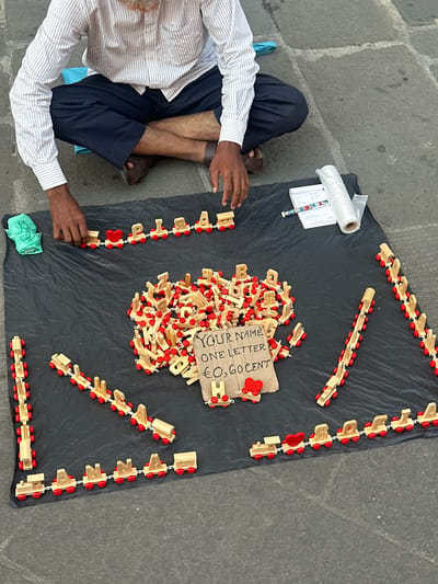 An overhead shot captures a street vendor or artisan seated on a stone pavement in Pisa, Italy, showcasing handcrafted items for sale on a black tarp. The person, mostly visible from the waist down, wears a white and blue striped shirt, dark trousers or shorts, and brown sandals. Their hands are positioned near the merchandise, suggesting they are either assembling the items or preparing them for a customer.

The primary products on display are numerous small, light-colored wooden blocks, each shaped like a letter of the alphabet and equipped with red wheels, resembling miniature train cars. These blocks are gathered in a large pile at the center of the tarp. Around this central pile, several chains of these letter-trains are neatly arranged in various lines and curves, with some featuring red heart-shaped blocks. One visible chain appears to spell "STRA" followed by a heart.

A handmade cardboard sign, placed amidst the wooden letters, clearly advertises the product: "YOUR NAME / ONE LETTER / € 0,60 CENT," indicating that personalized names can be formed from the letter-blocks for sixty cents per letter. On the left side of the display, a small notepad with some handwritten notes (including partially visible text like "Play"), a colorful pen, and a roll of clear plastic packaging are visible. The scene is brightly illuminated by natural daylight, and the ground is a textured grey stone pavement, characteristic of an urban setting.