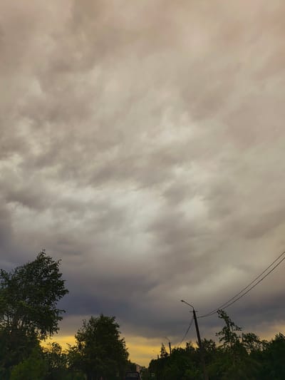 The image depicts an overcast sky above a street in Gremyachinsk, Russia. The sky is filled with thick, gray clouds, suggesting an impending storm. Below, trees and foliage line the street, partially obscuring the view of the road. Utility poles and power lines are visible against the cloudy sky. A warm, golden light is visible below the clouds on the horizon.