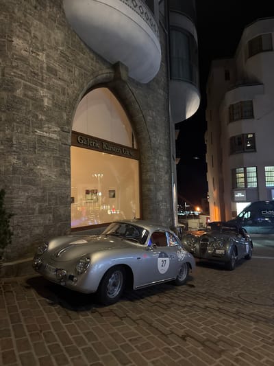 The image depicts a nighttime street scene in Sankt Moritz, Switzerland. Two vintage sports cars are parked on a brick-paved street, illuminated by artificial light sources.

The car in the foreground, positioned to the left, is a silver Porsche 356 coupe, bearing the competition number "27" on a white circular decal affixed to its driver's side door. Its front license plate reads "TG-194 YS".

Behind and to the right of the Porsche, a dark grey Jaguar XK roadster (likely an XK120 or XK140) is parked. Its front license plate displays "CT-811ZH".

To the left, a multi-story building constructed with dark, rough-hewn stone features a large, arched window opening. Above this window, the sign "Galerie Karsten Greve" is visible. The gallery interior, visible through the window, is brightly lit, reflecting various light sources from the street. A distinctive white, rounded architectural element projects from the upper portion of this building. A small evergreen shrub is visible at the base of the building on the far left.

To the right, across the street from the stone building, stands a lighter-colored, multi-story building with numerous windows and some small balconies. In the far background, a dark blue van with the word "Classic" partially visible on its side is parked. The sky is dark, indicating nighttime. No individuals are visible in the scene.