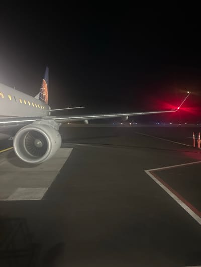 The image captures a static commercial aircraft, identified as a United Airlines airplane by its tail fin livery, positioned on an asphalt tarmac at night. The scene is located at Aspen Pitkin County Airport (ASE) in Aspen, United States. The aircraft's left side is prominently featured, displaying a section of the grey fuselage with illuminated passenger windows, the left wing, and a large turbofan engine. A bright red navigation light is active on the left wingtip, emitting a strong glow. The tarmac beneath the aircraft is dark asphalt, marked with distinct white and red painted lines forming an angle. Two orange traffic cones are visible on the far right side of the frame, near the aircraft's wingtip. Distant blue and red airport lights are faintly discernible on the horizon. No individuals are visible, and no active movements or interactions are depicted within the frame.