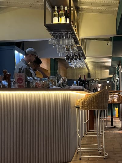 A rotated indoor shot captures a bar setting. A shelf holds wine bottles and glasses, while a counter displays jars and dispensers. Two people stand behind the bar. Wicker baskets hang above, and metallic chairs line the side. The wall and flooring feature linear patterns.