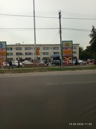 The image shows a street scene in front of a multi-story building, likely a government administrative building, in the city of Sokuluk, Kyrgyzstan. The building is light-colored with numerous windows and air conditioning units. In front of the building, there is a sidewalk and a landscaped area with planters. Several cars are parked in front of the building and along the street. Two tall signs with green and yellow designs are visible on the right side of the frame, one of which appears to be a speed limit sign indicating 40. A traffic light pole is also present. Two individuals are walking on the sidewalk in front of the building. The foreground shows a paved road, and a timestamp in the bottom right corner indicates the date "19.06.2026 11:52". The sky is overcast.