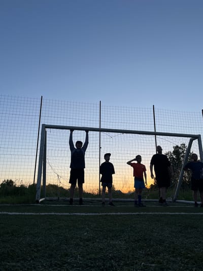 A group of five people, appearing to be young males, are silhouetted against a sunset on a sports field in Lebyazhye, Russia. They are standing in front of a soccer goal with a net. The sky is a gradient of clear blue to pale orange near the horizon. The grass field has white lines demarcating its boundaries. The individuals are dressed in casual athletic wear, including shorts and t-shirts. One person is hanging from the goal crossbar. The scene suggests an informal gathering or practice session at dusk.