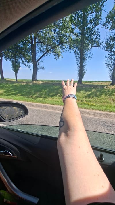 A person's arm is visible, extended out of a car window on a sunny day. The arm is adorned with two beaded bracelets, one pink and one light blue, and a watch. The person has a tattoo on their upper arm and a ring on their ring finger. The background shows a rural landscape with a road, a grassy embankment with scattered trees, and a clear blue sky. The scene appears to be in the vicinity of Дзяржынскі сельскі Савет, Belarus.