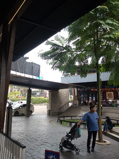 The scene depicts an outdoor plaza or entrance area to a commercial complex in Tân Kỳ Commune, Vietnam, under an overcast sky. The ground is paved with rectangular light grey blocks, appearing wet, possibly from rain.
In the foreground, a man, dressed in a blue t-shirt and dark trousers, stands with his left hand on the handle of a black baby stroller, identifiable by the "CHICCO" brand on its wheel. A light blue water bottle hangs from the stroller's handle. He faces slightly right, looking beyond the frame. To his left, a partial blue and red sign with a white circular symbol is visible on the pavement.
To the right, a large tree with abundant fern-like green foliage grows from an opening in the pavement. Further right, a person in dark clothing and a head covering sits on the lower steps of a concrete staircase with black railings, appearing to look at a device. These stairs lead up to a multi-story building featuring horizontal corrugated metal siding on its upper level and storefronts below, one with vertical red and white stripes and illuminated signs, including "ALL YOU CAN GELATO". A circular water feature with small jets is visible near the base of the tree and steps.
To the left, an extensive dark corrugated metal overhang extends from a building, supported by textured columns. Below a lower section of this overhang, a paved driveway or parking area is visible, with a white hatchback car parked or moving. A short, dense green hedge separates this area from a road. Above the lower overhang, large blank advertising hoardings are visible against the bright, overcast sky.
