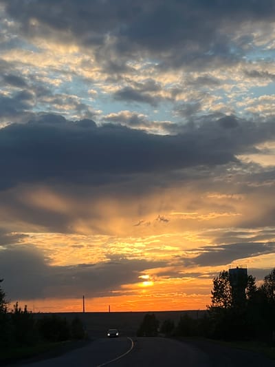 The image shows a sunset in Зыково, Russia. The sky is filled with clouds, with a mix of dark gray and light blue-white patches. The sun is setting on the horizon, casting a golden orange glow across the landscape. There is a road with a car on the right side of the image, with trees and vegetation bordering the road. In the distance, thin vertical structures are visible on the horizon.