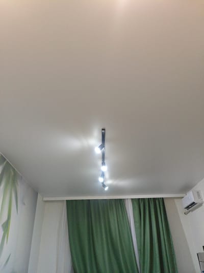 The image shows a close-up view of a white ceiling with a black track light fixture holding several small, bright bulbs. The lights are illuminated, casting a glow on the ceiling. To the left, a portion of a wall with a mural of large, pale green leaves is visible. To the right of the track light, green curtains are drawn, partially obscuring the wall. A white air conditioning unit is mounted on the wall above the curtains. The scene appears to be an indoor residential or commercial space. There is no discernible information in the image that would allow for the specific location in the city of Kalach-na-Donu, Russia.