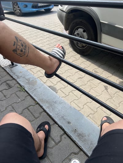 The image shows a person's legs and feet, wearing flip-flops. One leg has a tattoo of a cat. The person is sitting or standing outdoors, with a metal railing in the foreground. In the background, there is a blue car and a white van, parked on a paved surface. The scene appears to be in an urban or suburban environment, possibly a parking lot or street in Michałowice, Poland. The ground is covered with interlocking paving stones.