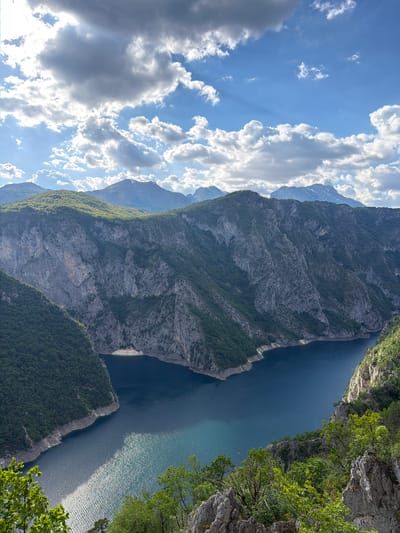 The photograph depicts a high-angle, outdoor landscape in Plužine, Montenegro. A deep blue body of water, likely a lake or river, stretches vertically through the center of the frame. On either side of the water are steep, rocky cliffs covered with sparse vegetation. The cliff on the left appears to be significantly taller and stretches towards the left side of the frame, partially obscuring mountains behind it. The cliff on the right is closer to the viewer and has trees growing near its edge. Above the cliffs and mountains, the sky is visible with scattered white clouds.