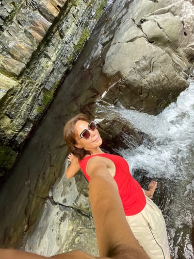 A woman with sunglasses and a red top is relaxing in a natural pool formed by a waterfall. She is lying on her back with her head tilted up, looking towards the camera. Her legs are extended, and one foot is visible submerged in the clear water. The surrounding rocks are wet and appear to be layered, with some patches of green moss growing on them. The waterfall is cascading down the rocks, creating a foamy white water effect. The overall scene suggests a peaceful and refreshing moment in nature, likely during a warm day. The image appears to be a selfie, taken from a low angle. No text is visible in the image. The location context provided is Bolshoy Sochi, Russia.