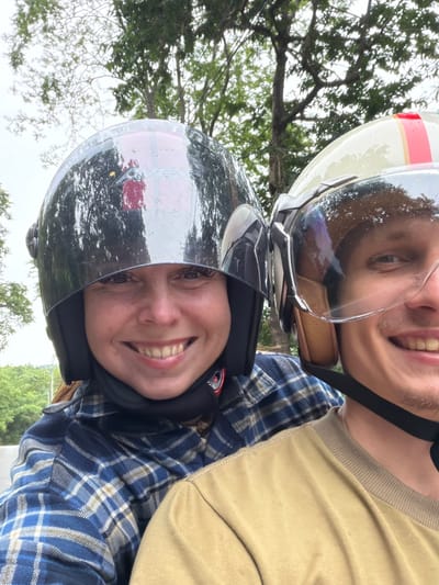 Two individuals are wearing helmets and riding on a motorbike. The person in the foreground, on the left, is a woman with brown hair, wearing a blue and white plaid shirt and a black helmet with a reflective visor. She is smiling broadly. The person behind her, on the right, is a man with short hair, wearing a tan shirt and a cream-colored helmet with a clear visor. He is also smiling. They appear to be on a road with trees and foliage in the background, suggesting a rural or natural setting. The weather appears overcast. The image was taken in Samoeng Tai, Thailand.