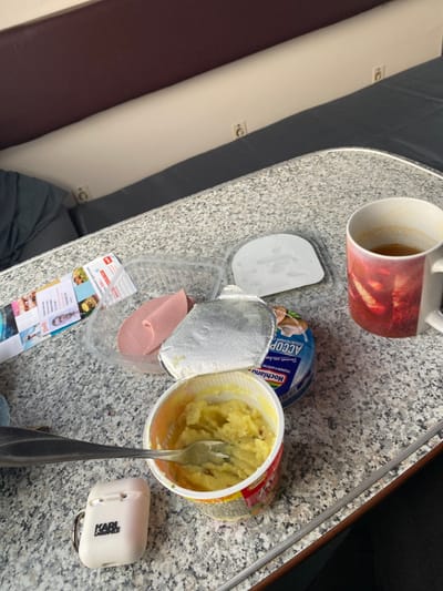 The image displays a meal setup on a speckled gray laminate table, likely within a train compartment. The immediate background features a light beige wall panel with two visible power outlets and a dark gray upholstered section below it. A maroon upholstered bench seat runs along the top edge of the frame.

On the table, from left to right, are: a folded leaflet with colorful imagery and partially visible Russian text; an open clear plastic container holding two slices of pink processed meat; an open white Karl Lagerfeld branded AirPods case; an open white plastic cup containing mashed potatoes with a silver fork inserted into them; a blue Hochland processed cheese container, partially open with its foil lid peeled back, displaying Russian text including "ПЛАВЛЕНЫЙ" and "АССОРТИ"; and a white ceramic mug with a red and orange abstract design, holding a partially consumed dark beverage. A small, flat white plastic piece is situated near the mug.

No individuals are visible. The objects suggest a recent or ongoing meal. The Russian text on packaging indicates a Russian-speaking environment. While the overall setting is consistent with a train car in Russia, the specific location within the city of Yar, Russia, cannot be determined from the visual information provided.