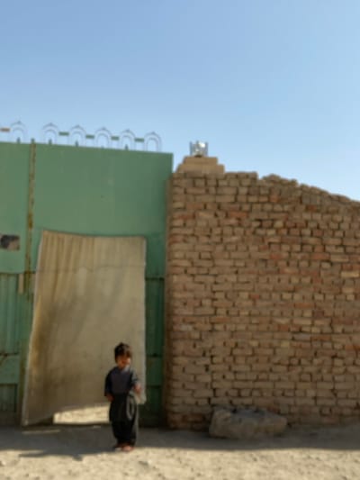A low-angle shot presents a child standing in front of a green-gated building, adjacent to a brick wall. The wall appears to be older with some bricks missing near the top. Above the green building, a line of curved barbed wire stretches across the top. The sky in the background is clear and blue. The perspective is slightly off-kilter, causing a tilted appearance.